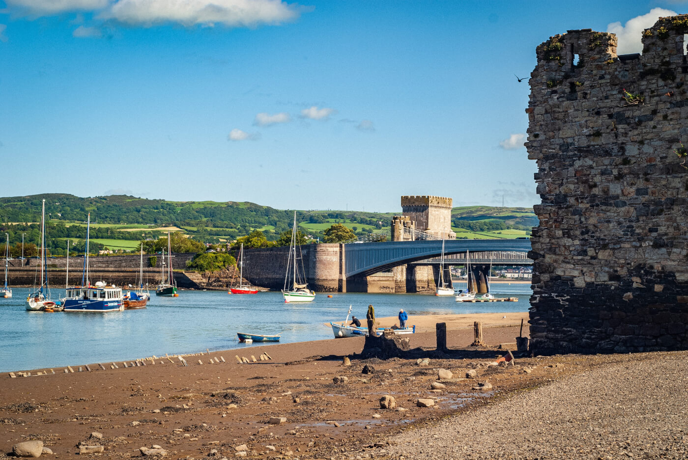 Image of a beach with views of docked boats, Conwy Bridge and part of the castle.